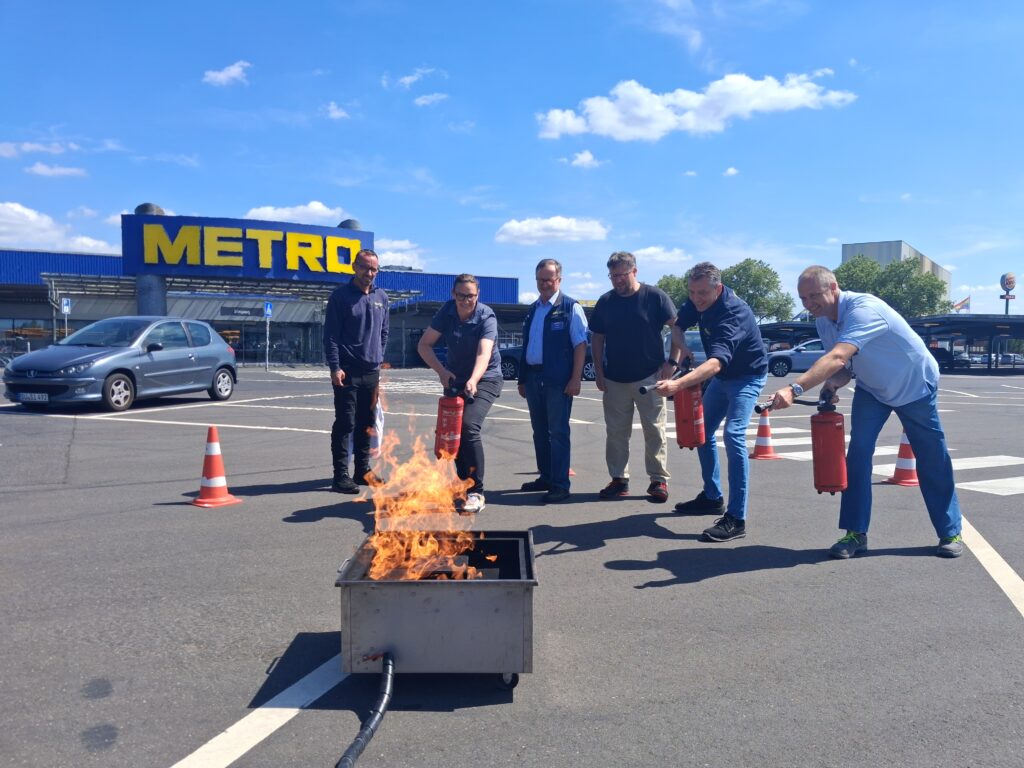 Brandschutzhelfer bei einer praktischen Feuerlöschübung auf dem Parkplatz eines Unternehmens mit einem gasbefeuerten Übungsbrand und Feuerlöschern.